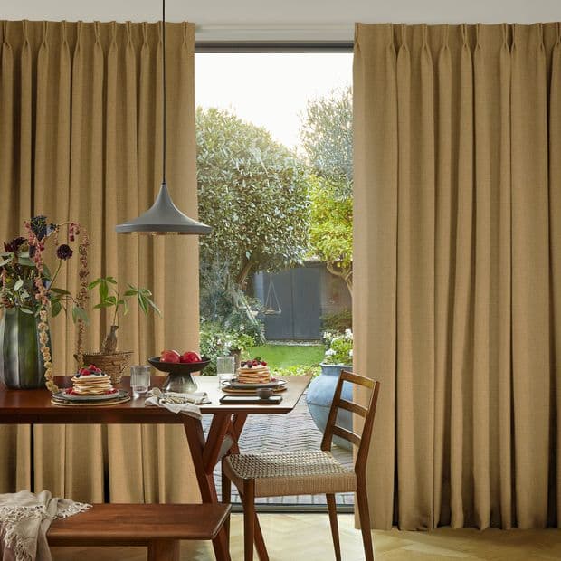 Floor-length beige pleated curtains part at center, framing a glass sliding door; curtains draw back, letting daylight filter into a dining area with wooden table, chairs, pendant light and garden view.