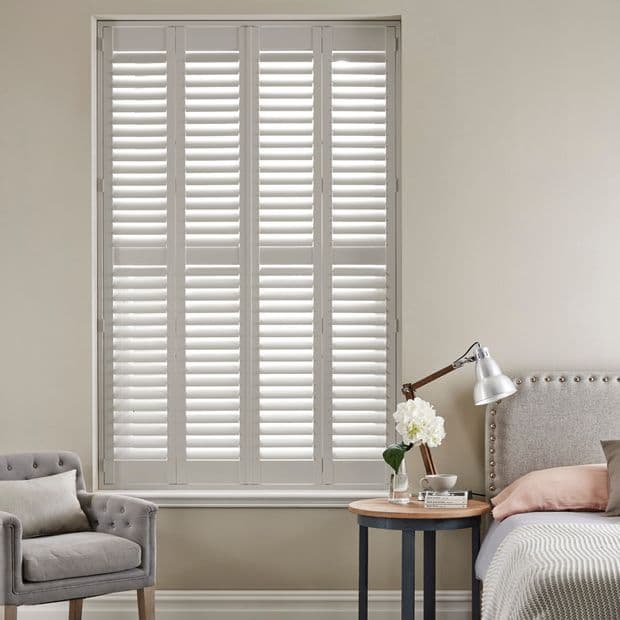 White full-height louvered plantation shutters covering a tall window, slats slightly tilted filtering soft daylight; bedroom setting with upholstered bed, bedside table and lamp, neutral walls and armchair.
