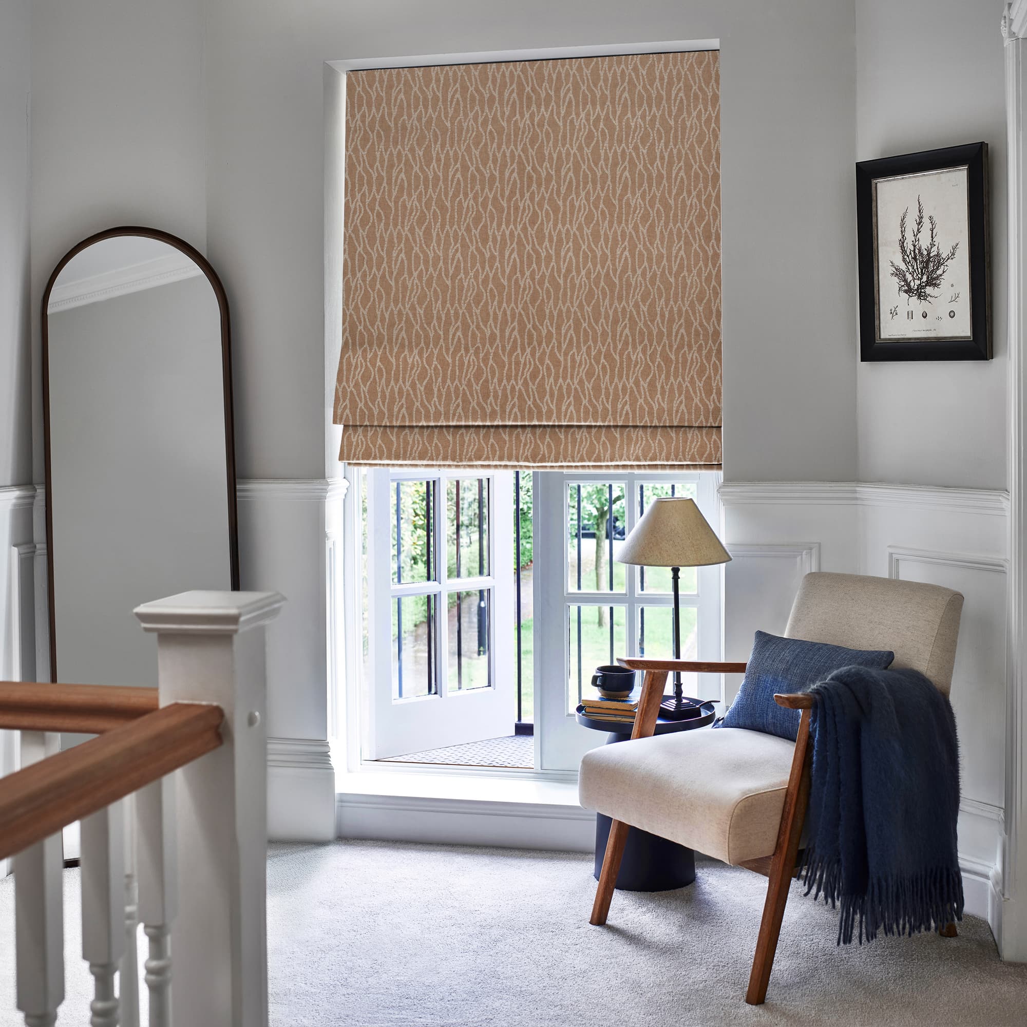 Patterned beige Roman blind, lowered and slightly folded at the bottom, filtering daylight over a ground-floor casement window opening to a garden, in a lit landing with armchair, lamp, mirror.