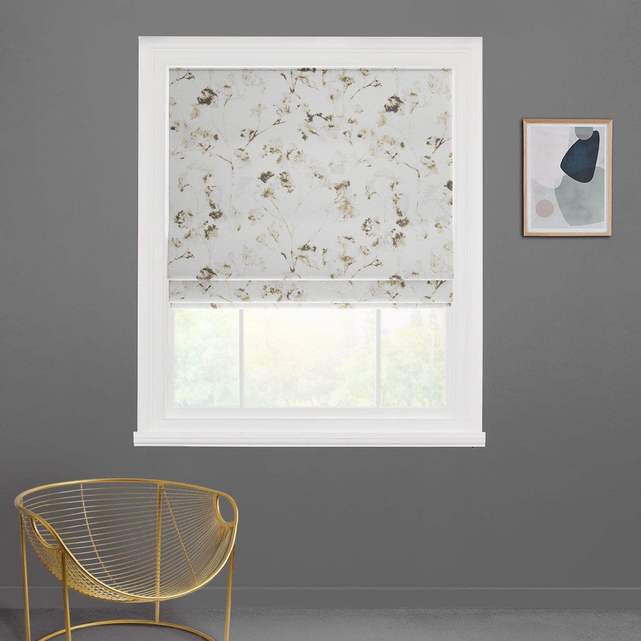 A white Roman blind with pale gold floral pattern, lowered and filtering daylight, in a white window frame on a gray wall beside an abstract print and a gold chair.