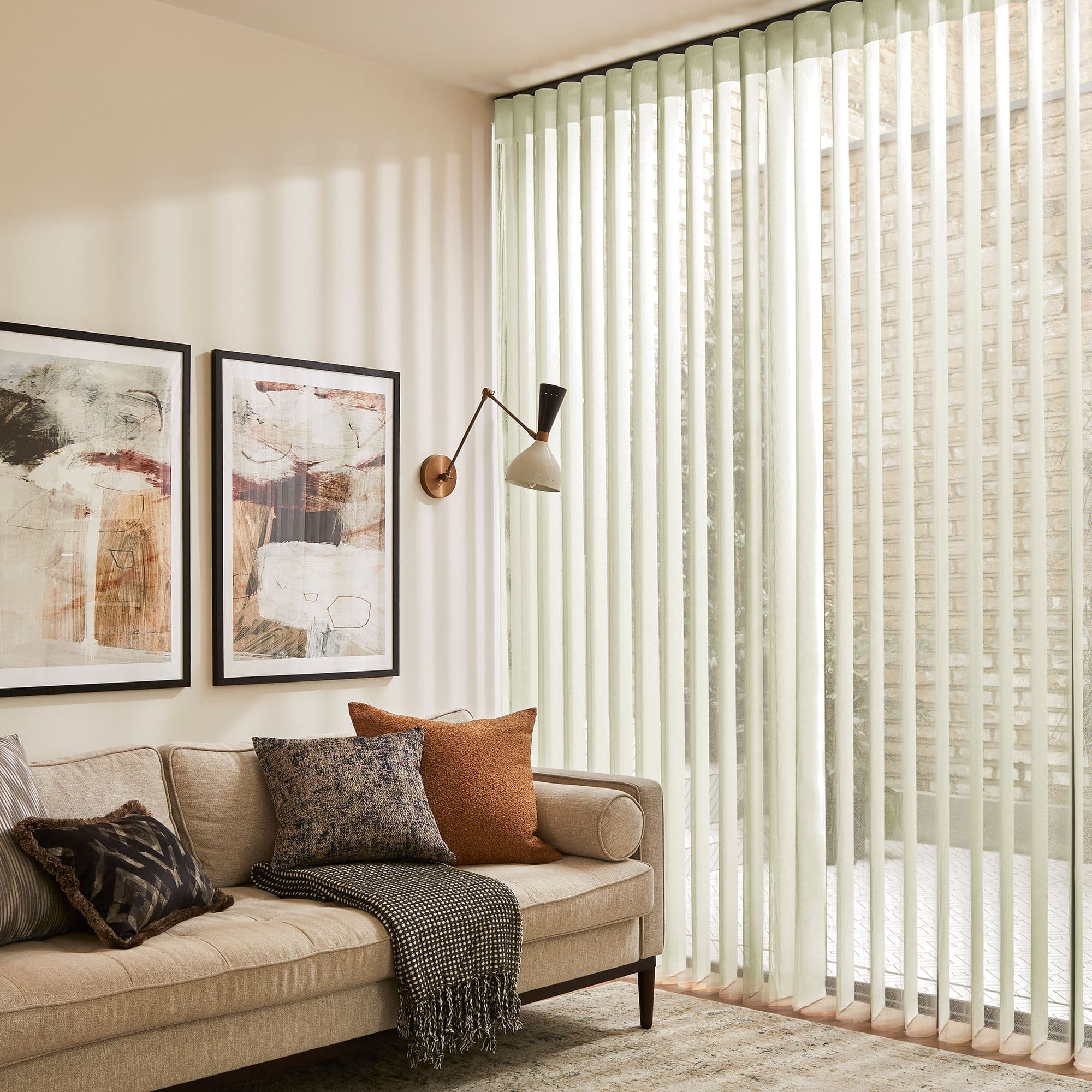 Pale fabric vertical blinds cover a floorâ€‘toâ€‘ceiling sliding glass door, slats angled to filter soft daylight, casting stripes; cozy living room with beige sofa, cushions, wall lamp and framed art.
