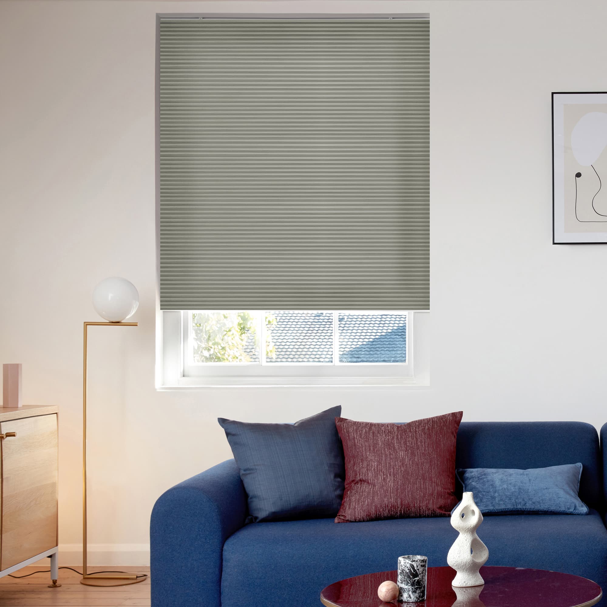 Gray pleated cellular blind, fully lowered across the window with fine horizontal ribs; blocking and softly filtering daylight; small living room setting with a blue sofa, cushions, floor lamp, wall art.