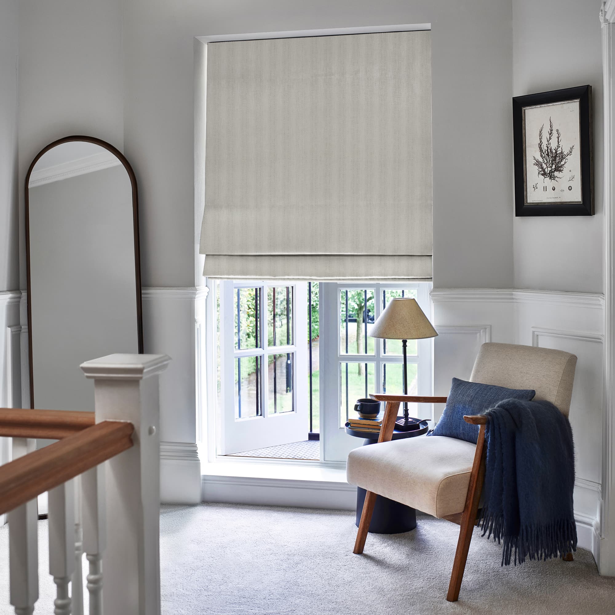 Beige fabric Roman blind, lowered with soft horizontal folds, filtering daylight; in a carpeted upstairs landing reading nook with armchair, navy throw, side table lamp, tall mirror, and garden view.