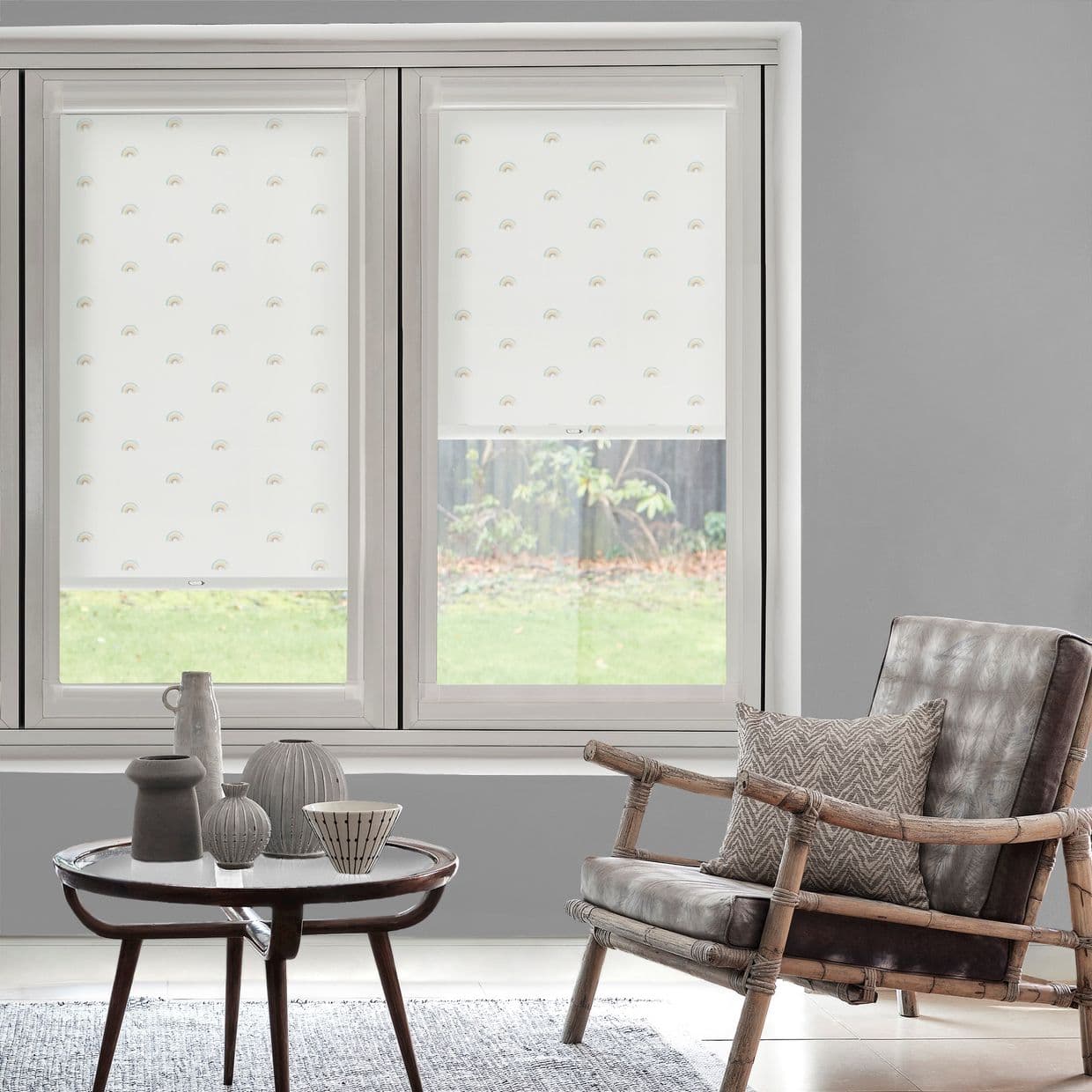 White roller blinds with tiny rainbow motifs, fitted inside a twoâ€‘pane window â€” left fully lowered, right partially raised, softly filtering daylight; living room with gray walls, rattan chair and coffee table.