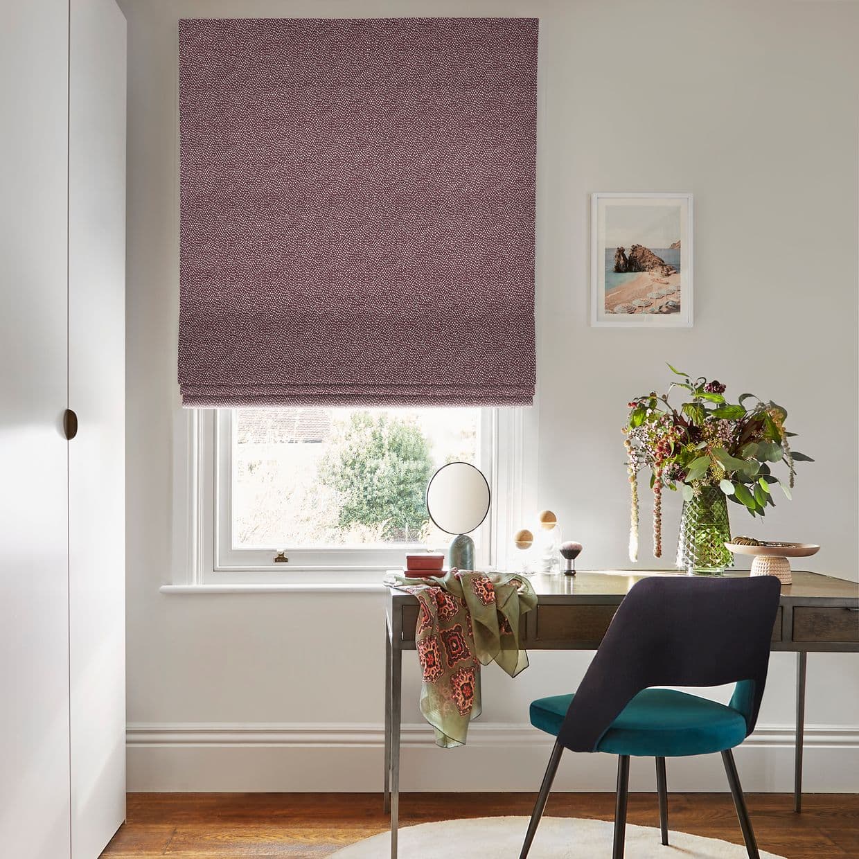 A patterned maroon Roman blind lowered over a window, mostly closed and filtering daylight; small desk with mirror, teal chair, vase of flowers and framed print in a bright room.