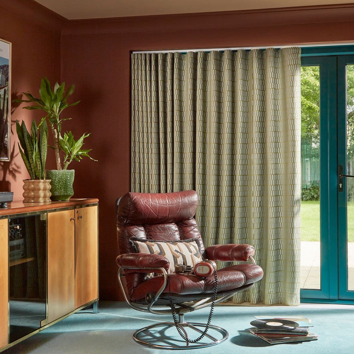 Pleated geometric beige-green curtains covering a teal-framed sliding glass door, drawn closed and softly filtering daylight; cozy living room with burgundy leather recliner, wooden cabinet, potted plants, records on carpet.