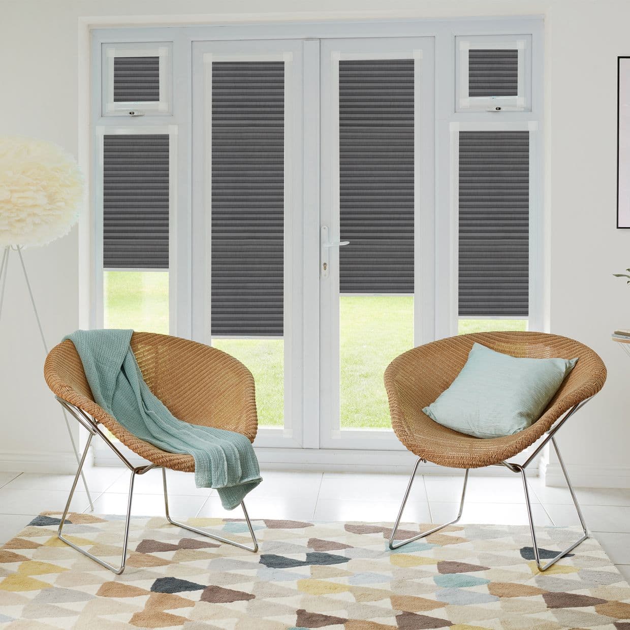 Pleated dark-grey cellular blinds covering tall French doors and upper transom windows, lowered to partially block light, in a bright modern living area with wicker chairs and patterned rug.