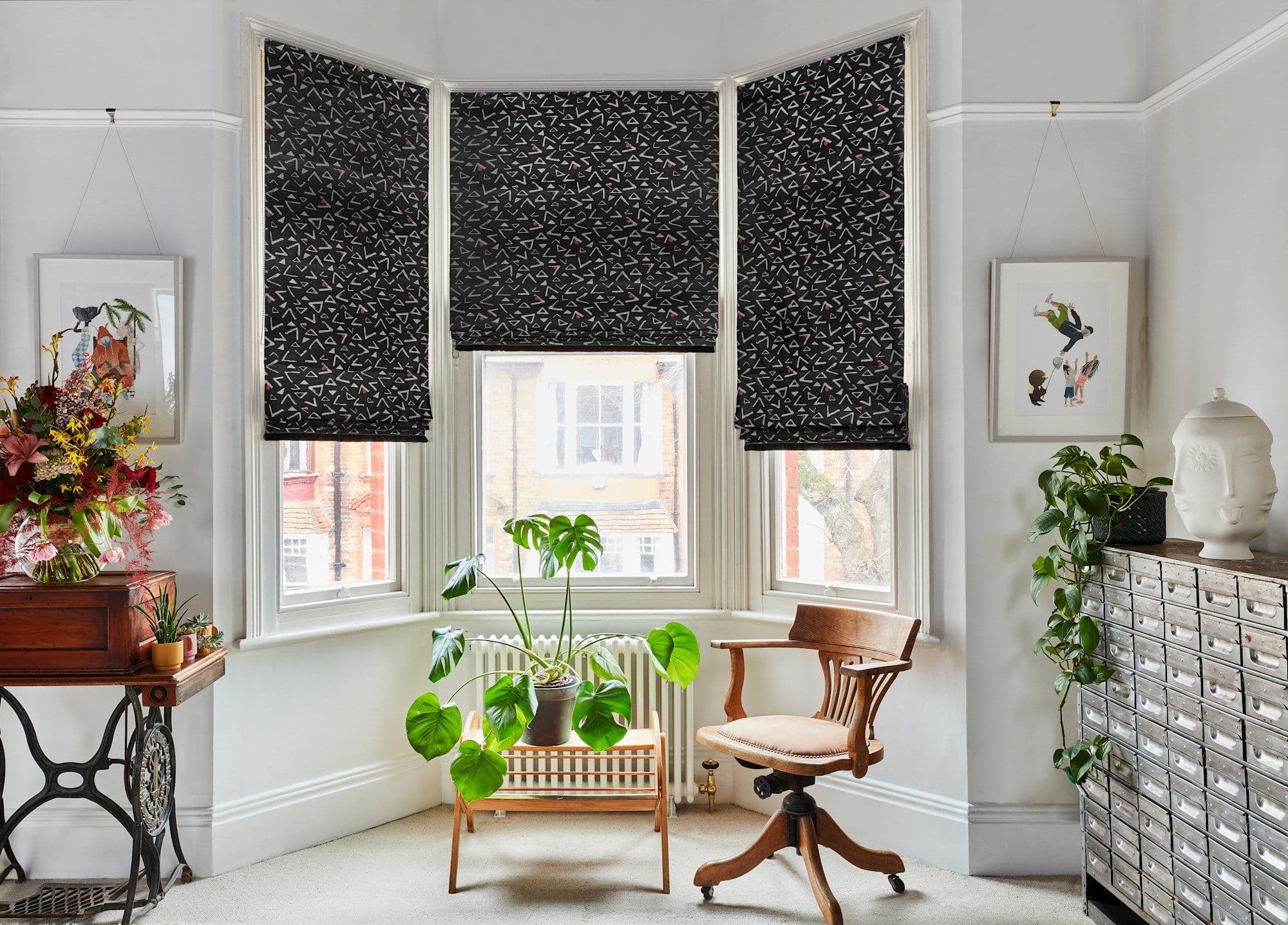 Three patterned black Roman blinds across a bay window, mostly lowered and filtering daylight; central blind slightly raised; room contains a wooden swivel chair, potted plants, and a metal storage cabinet.