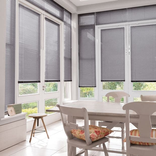 Grey horizontal venetian blinds, lowered over tall bay windows with slats mostly closed and bottoms slightly raised, filtering daylight into a bright white dining nook with table, chairs, floral cushions.