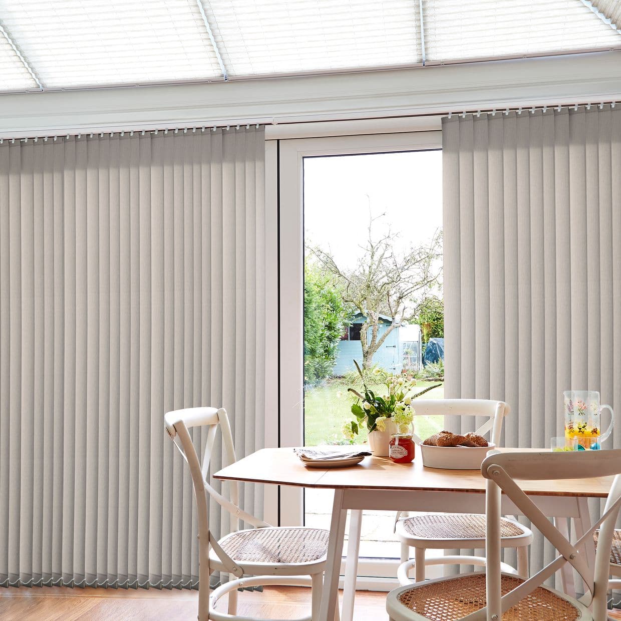 Beige vertical blinds mounted across a sliding patio door, mostly closed with a narrow central opening, filtering daylight and revealing a garden, in a dining area with table, chairs, vase.