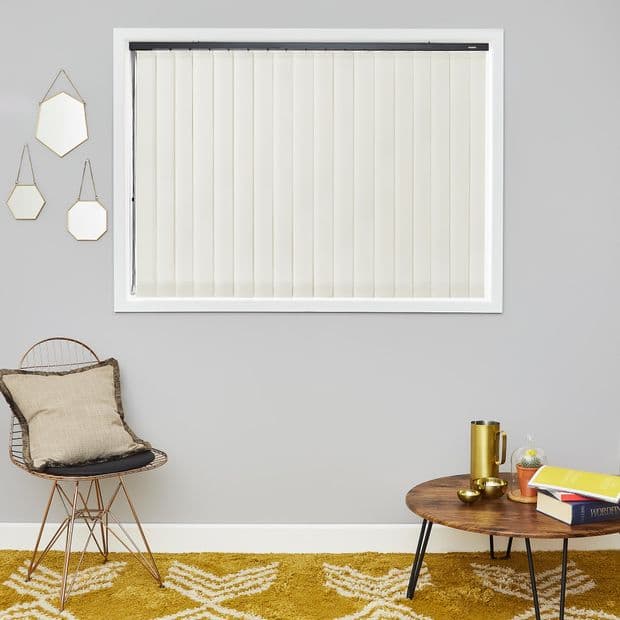 Cream vertical slat blinds, fully closed inside a white-framed window, gently filtering daylight; minimalist living room with gray walls, wire rattan chair, mustard patterned rug and round wooden coffee table.