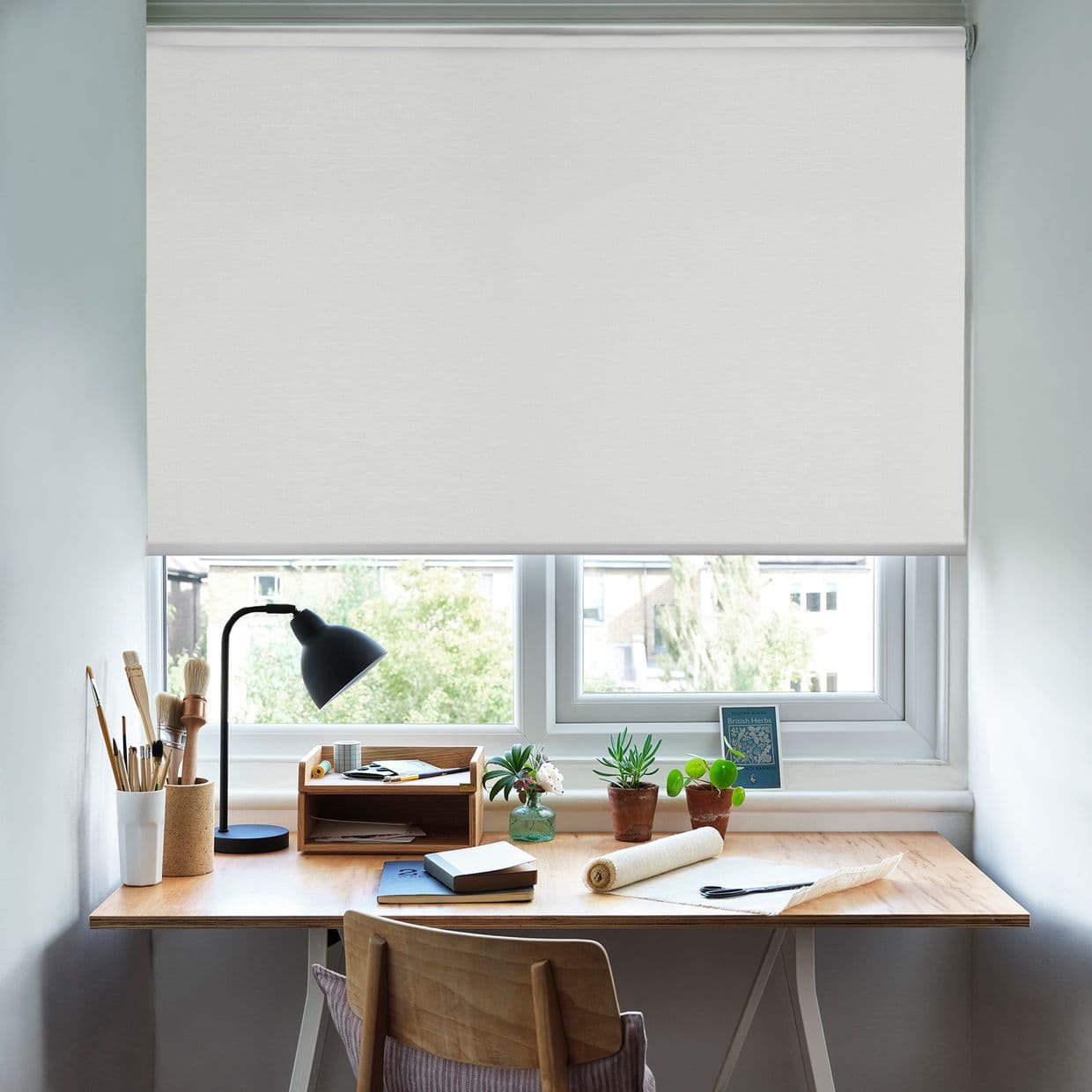 White roller blind, smooth fabric, almost fully lowered over the window; filtering soft daylight onto a tidy wooden desk with lamp, potted plants, art supplies and a suburban view.