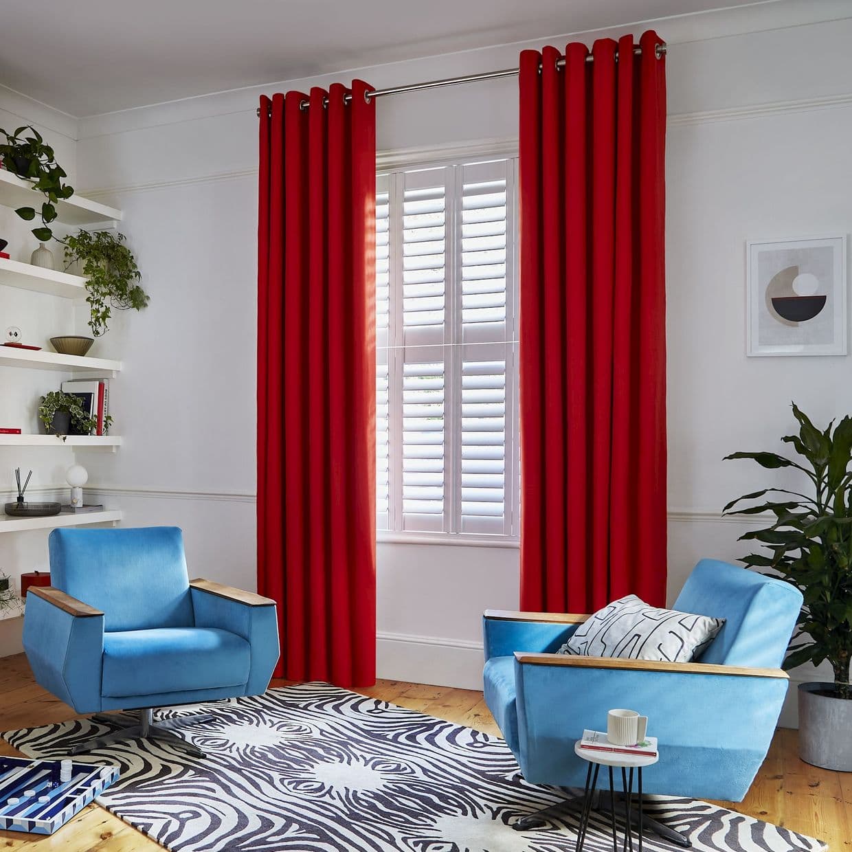 Red grommet-top curtains flank a window with white plantation shutters; curtains drawn open, shutters tilted to diffuse daylight. Context: bright living room with blue armchairs and a zebra-pattern rug.