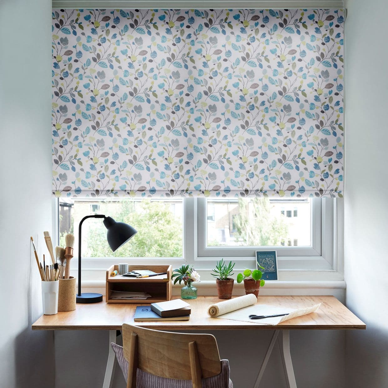 Floral roller blind lowered over two-pane window, white fabric patterned with blue, green, grey leaves; filtering soft daylight into a home study with wooden desk, lamp, art supplies.