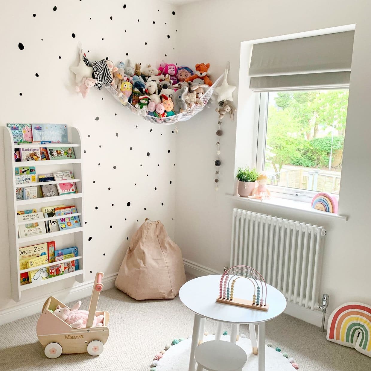 Grey Roman fabric blind (folded tiers) partially lowered, filtering daylight through the window; bright children's nursery with white polka-dot walls, stuffed-animal hammock, bookshelf, beanbag, small table, garden view.

Text found: Maeva
Text found: Dear Zoo