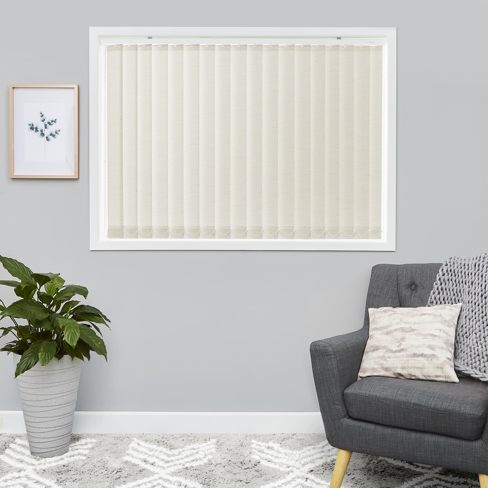 Vertical beige blinds mounted inside a window frame, fully closed and gently diffusing daylight; in a gray-walled living room with a gray armchair, knitted throw, potted plant and rug.