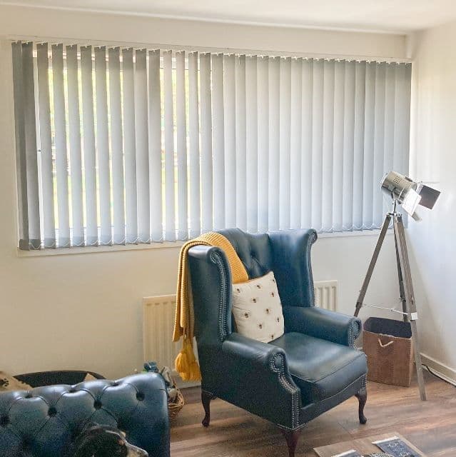 Vertical fabric blinds spanning a wide window, closed and softly filtering daylight; traditional blue leather wingback chair with throw and pillow faces into a bright, minimally furnished living room.