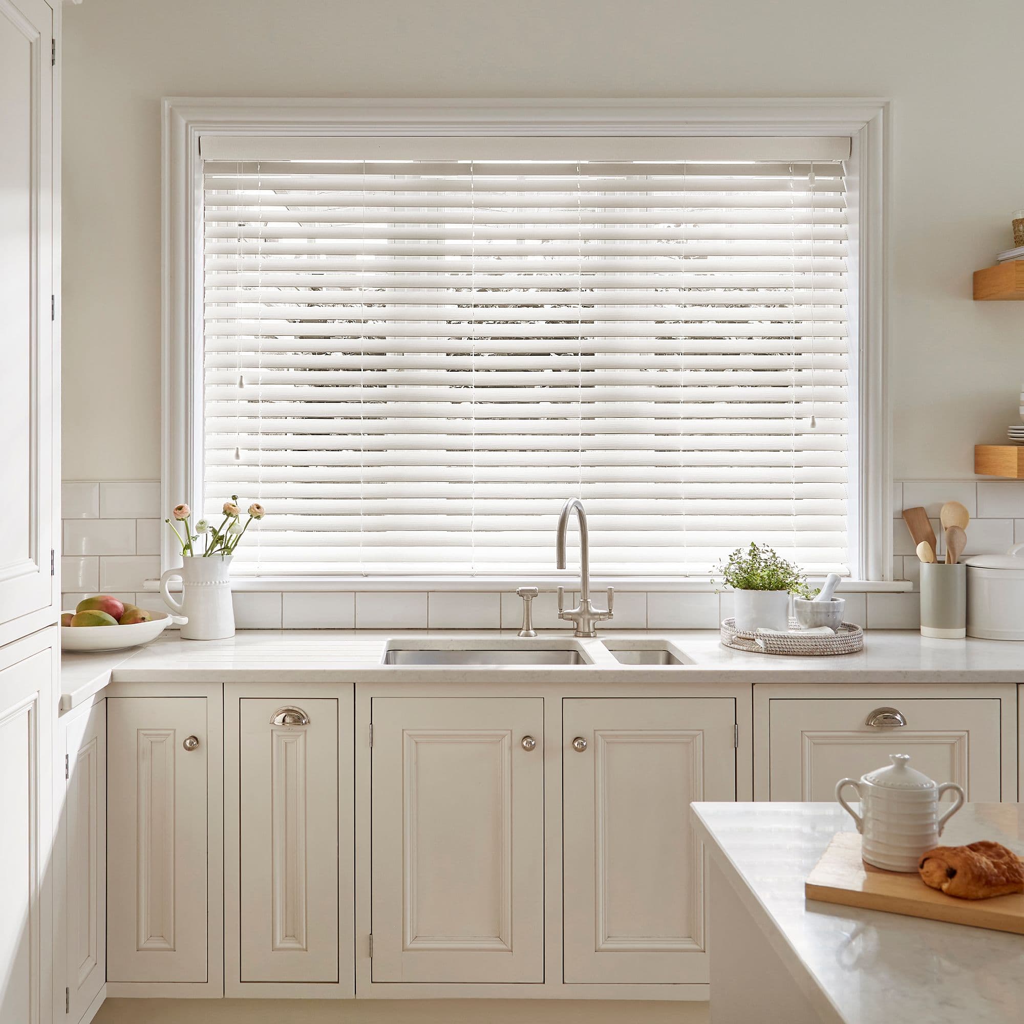 White horizontal Venetian blinds fully lowered across a kitchen window; slats slightly tilted filtering soft daylight into an airy cream-toned kitchen with marble counters, farmhouse sink, cabinets and simple decor.