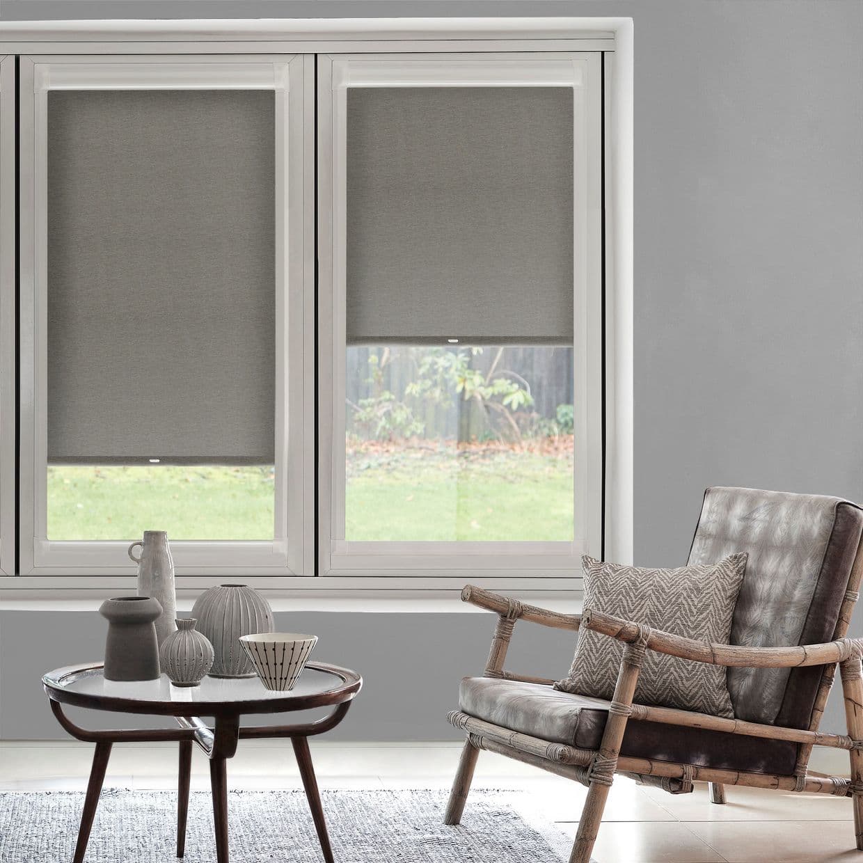 Two grey fabric roller blinds lowered mid-height across a double window, filtering daylight; a bamboo-framed leather armchair and round table with ceramic vases sit in a neutral living room.