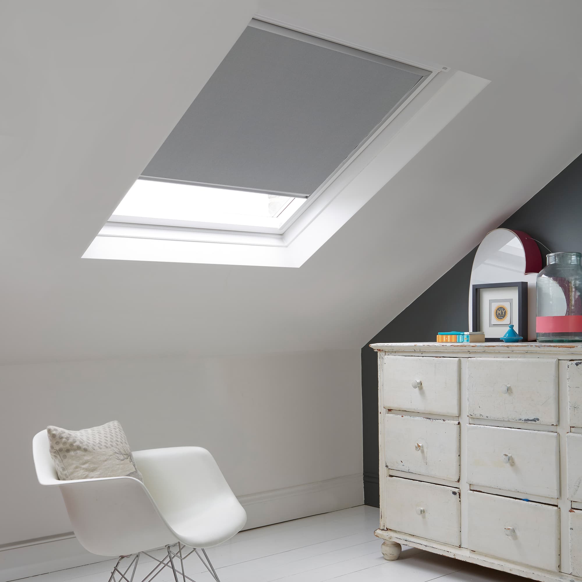 Room with white eaves wall, dark blue-grey wall to right with a shabby chic white drawer unit, a contemporary white chair, white painted floorboards and a skylight blind in steel.