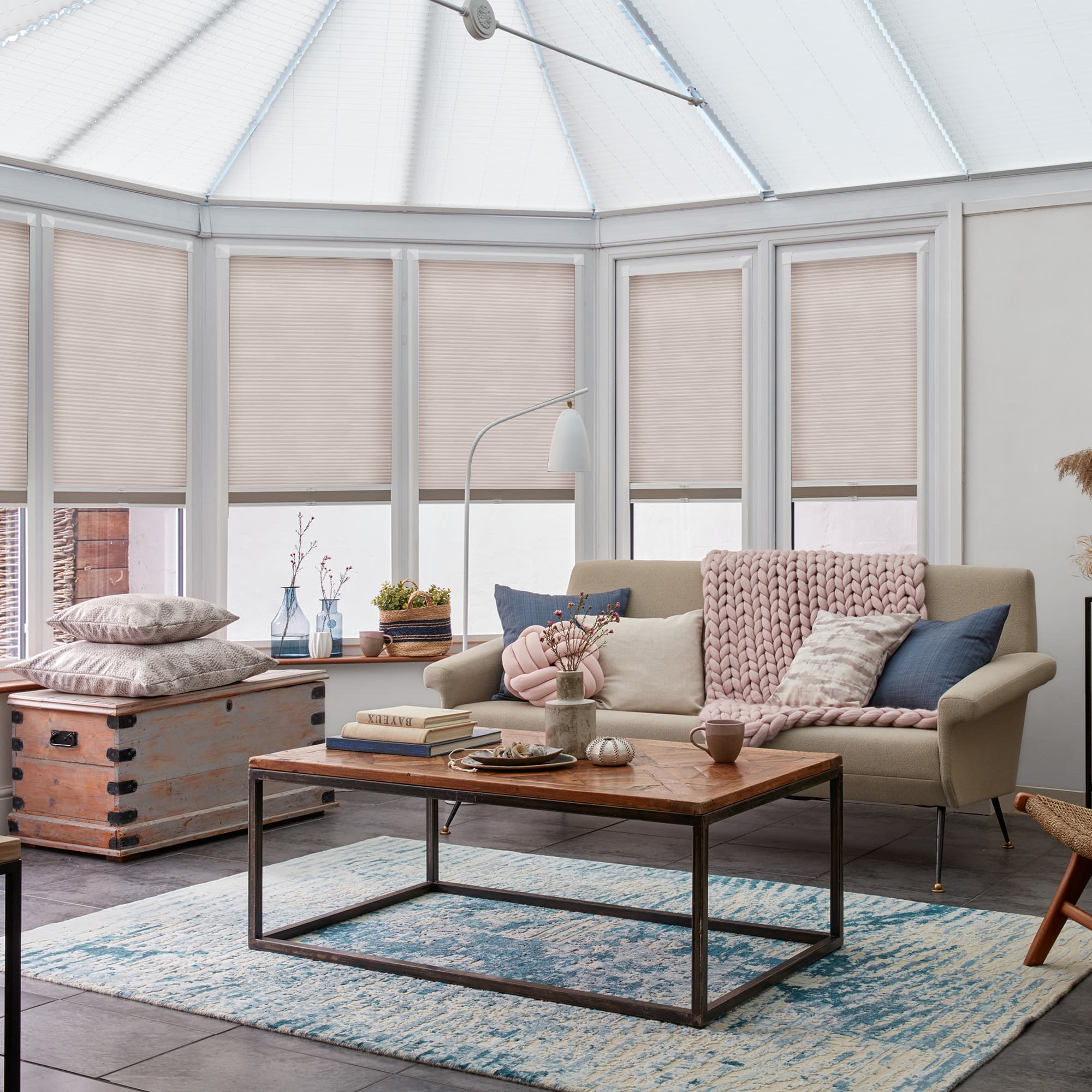Pleated beige cellular blinds, lowered across bay windows and conservatory panels, closed and diffusing daylight; conservatory living area with neutral sofa, chunky pink throw, wooden coffee table and blue rug.