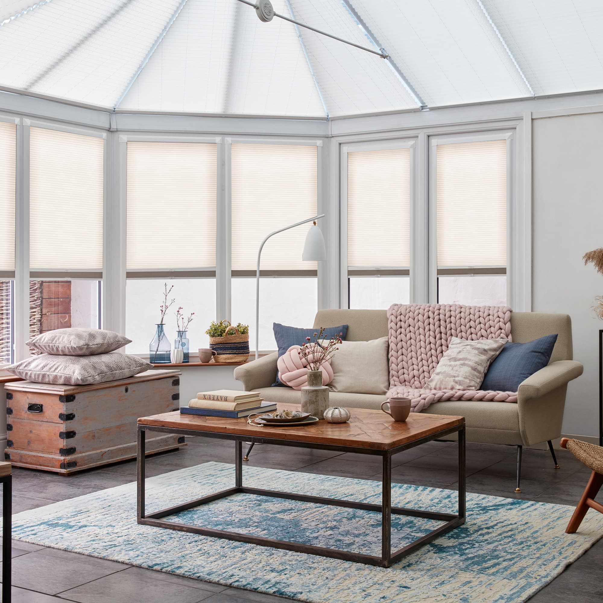 Pleated cellular blinds, fully lowered on bay windows with lightâ€‘filtering opaque fabric and narrow horizontal pleats; filtering soft daylight into a conservatoryâ€‘style living room with sofa, coffee table, rug.