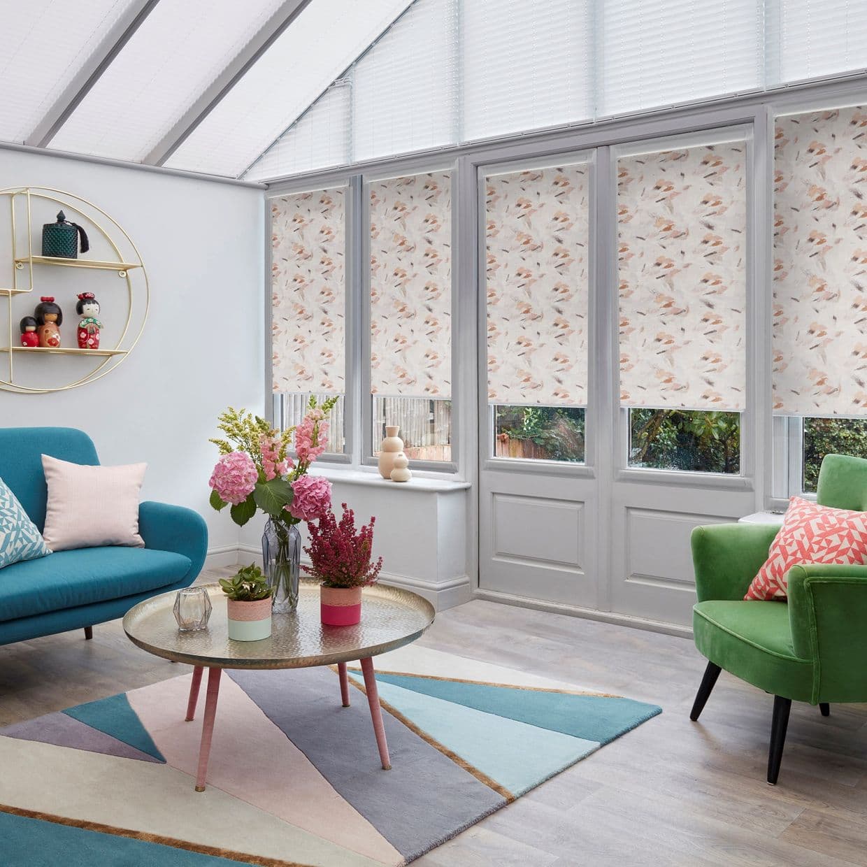 Patterned floral roller blinds, lowered over tall conservatory windows and doors, partially blocking and softly diffusing daylight; modern sunroom with teal sofa, green armchair, round coffee table and flowers.