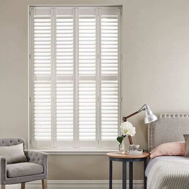 White plantation shutters covering a tall window, horizontal slats angled to filter bright daylight, in a softly lit bedroom corner with upholstered bed, side table, lamp and armchair.