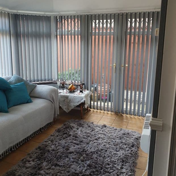 Grey vertical blinds cover bay windows and patio doors, slats partially open filtering daylight; conservatory with tiled floor, sofa with teal cushions, lace-covered side table and a grey shag rug.