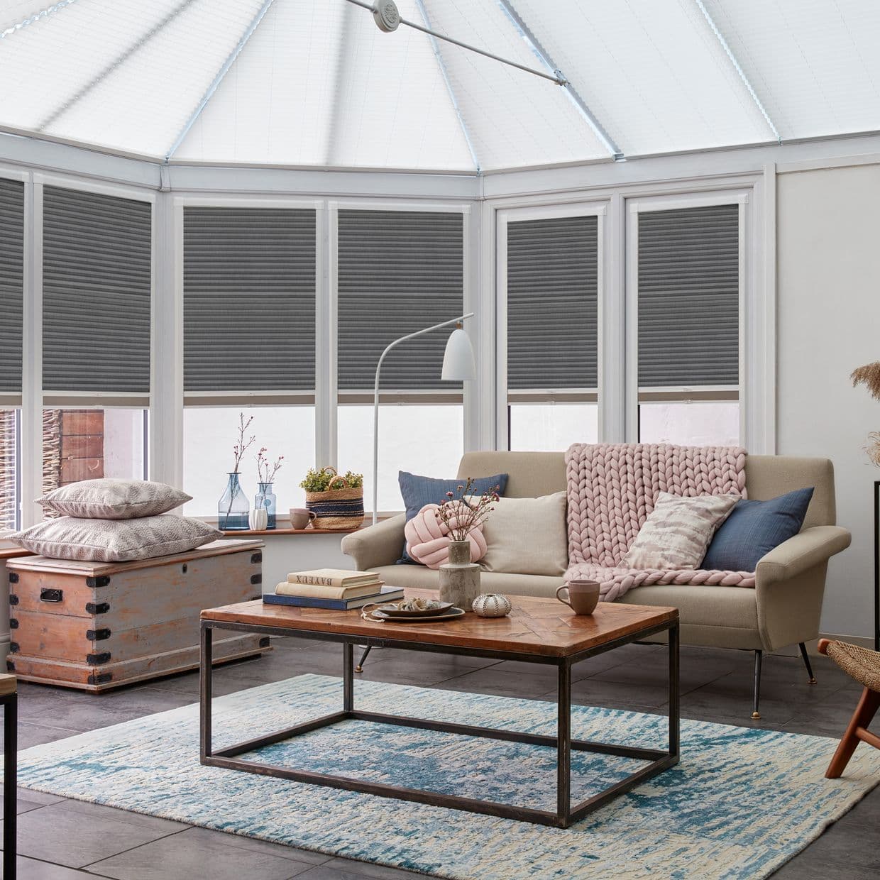 Grey cellular pleated blinds, lowered across tall conservatory windows, blocking and filtering daylight; in a bright conservatory-style living room with a beige sofa, wooden coffee table, blue rug, and soft textiles.