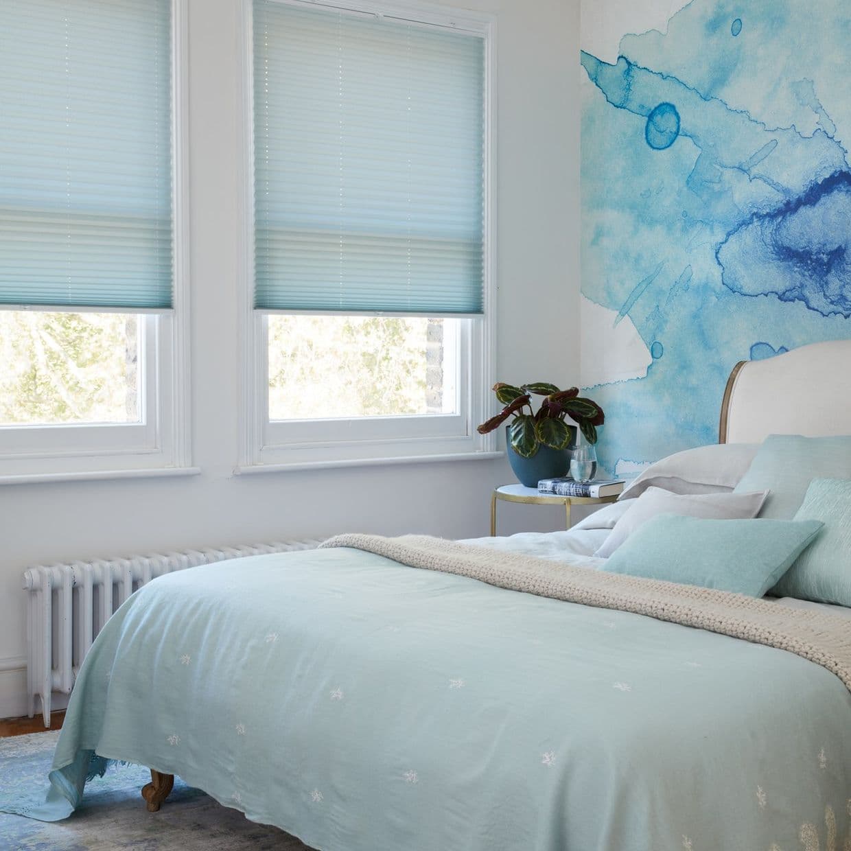 Light blue pleated cellular blinds lowered halfway, filtering soft daylight into a pastel bedroom with a pale aqua bed, watercolor blue accent wall, and a potted plant on a bedside table.