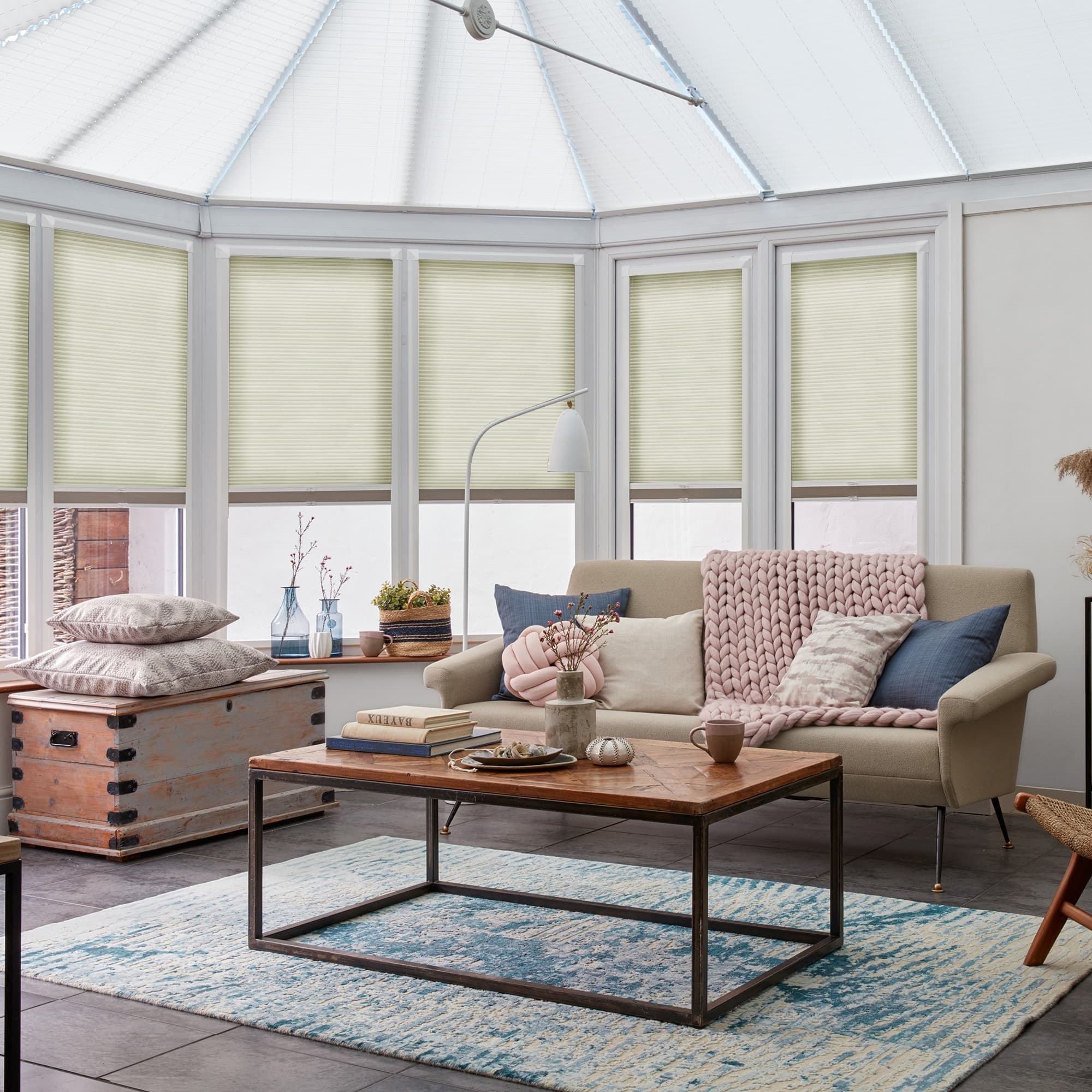 Cream cellular blinds lowered on conservatory windows and roof panels, leaving lower sashes exposed; filtering soft daylight into a bright sitting area with beige sofa, coffee table and blue rug.