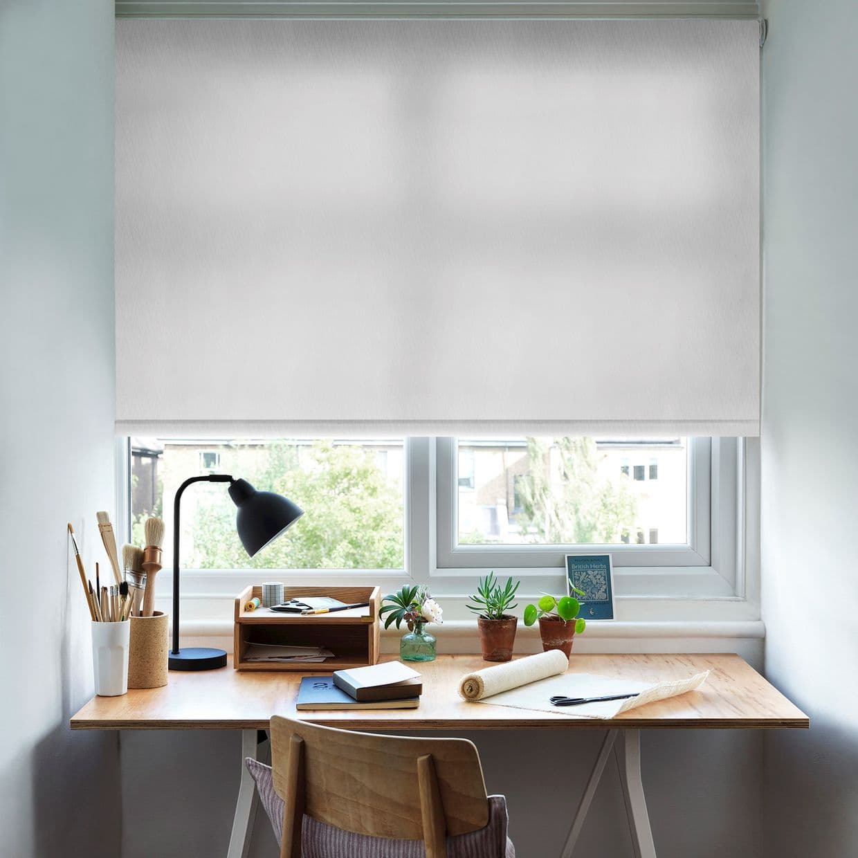 White roller blind, lowered nearly fully, translucent fabric diffusing soft daylight; over a home workspace with a wooden desk, black lamp, paintbrushes, notebooks, potted succulents and a leafy window view.