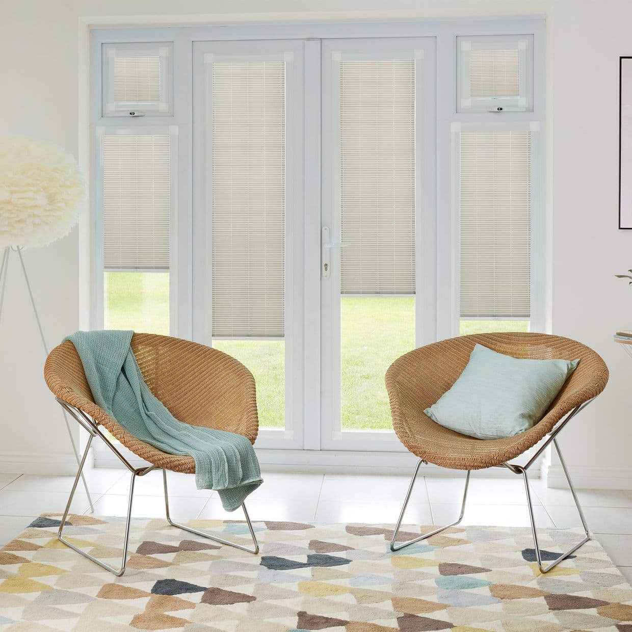 White Venetian-style blinds covering French doors and flanking sidelights, mostly closed with lower edges slightly raised to reveal lawn; filtering soft daylight into a neutral living room with wicker chairs.