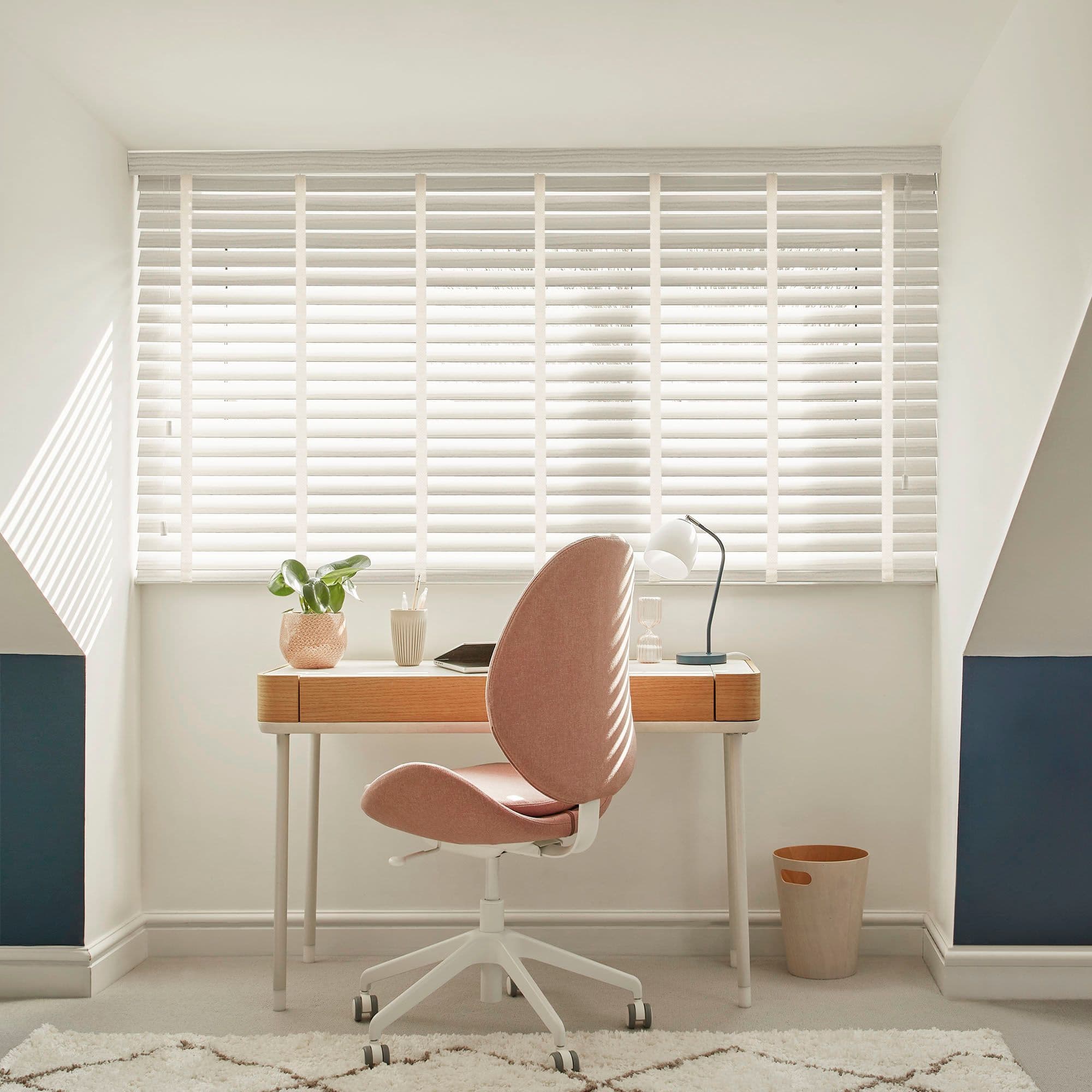 White horizontal blinds span the window, closed with thin slats and pull cord, filtering daylight and casting striped shadows over a wooden desk and pink swivel chair in an attic study.