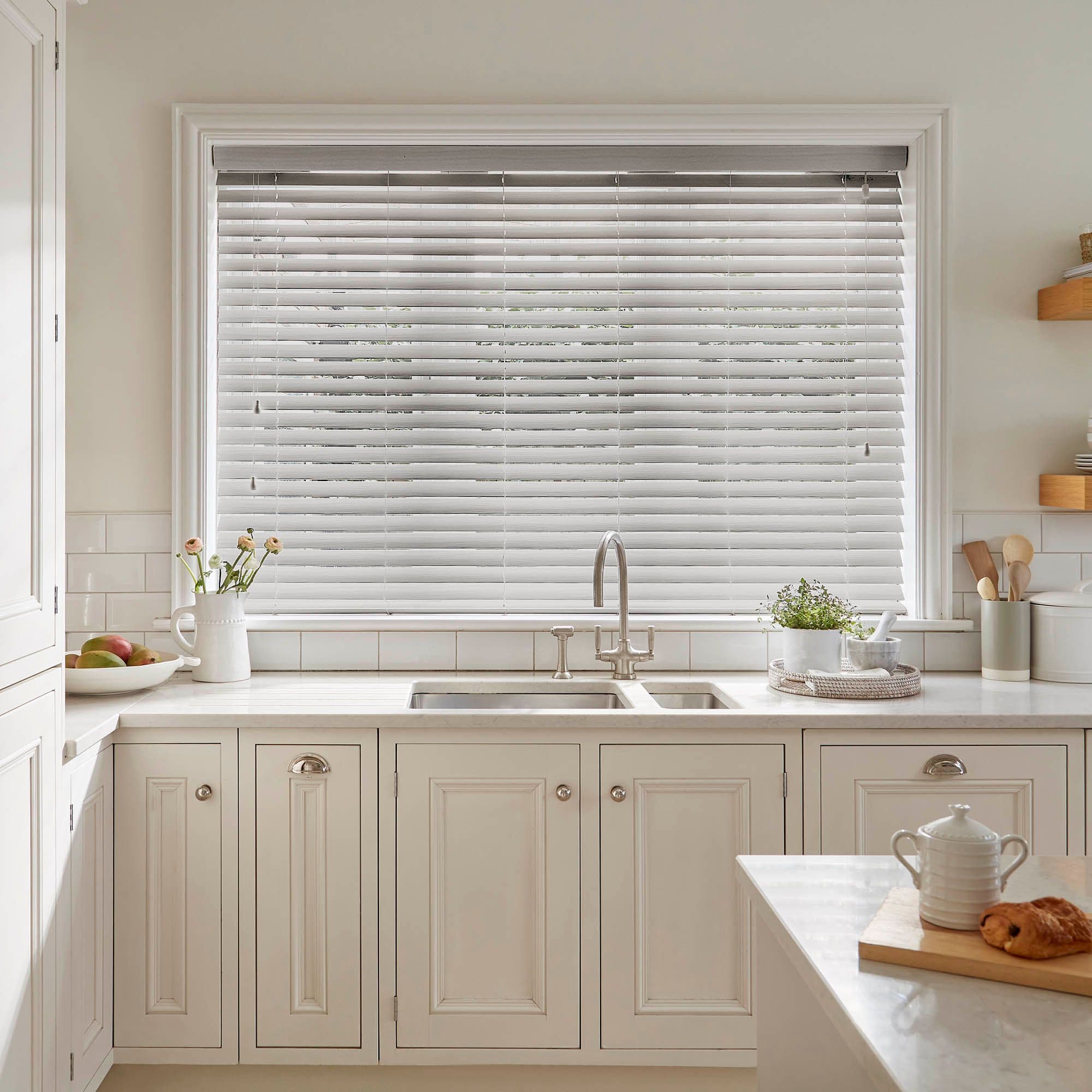 White horizontal Venetian blinds fully covering a large window, slats slightly tilted to filter soft daylight; above a cream kitchen sink and marble countertop with faucet, plants, and utensils.