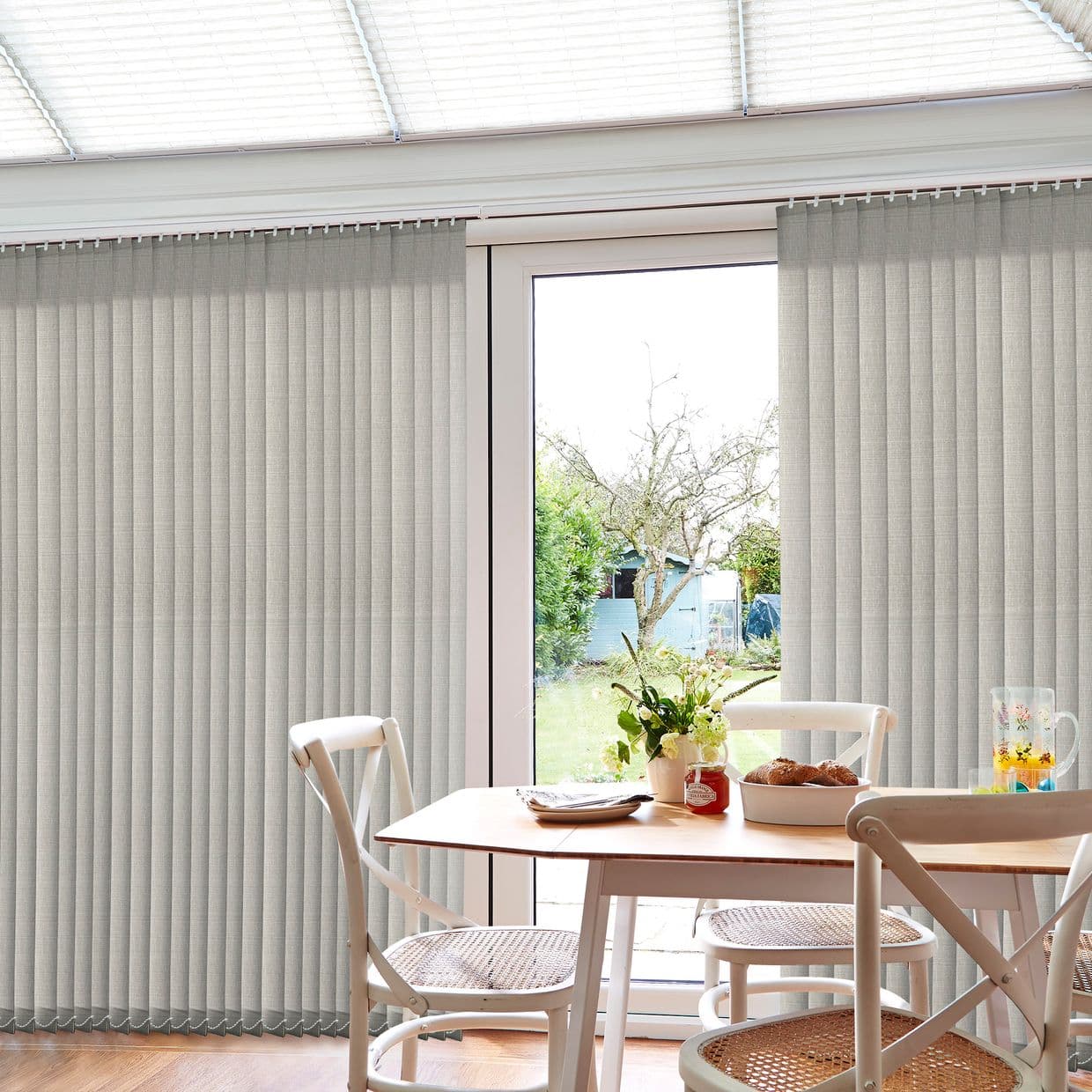 Beige fabric vertical blinds with pleated slats on a ceiling track, parted at center to reveal sliding glass door; dining set in foreground, soft daylight, backyard tree and shed.