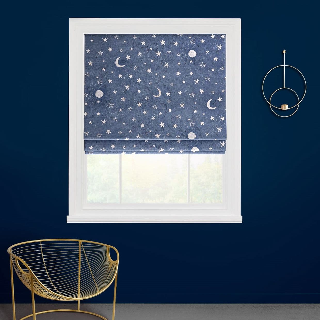 A navy-blue roman shade with white moon-and-star print, lowered to cover the upper window and filter daylight; set in a white-trimmed sash on a deep-blue wall with a gold chair and sconce.
