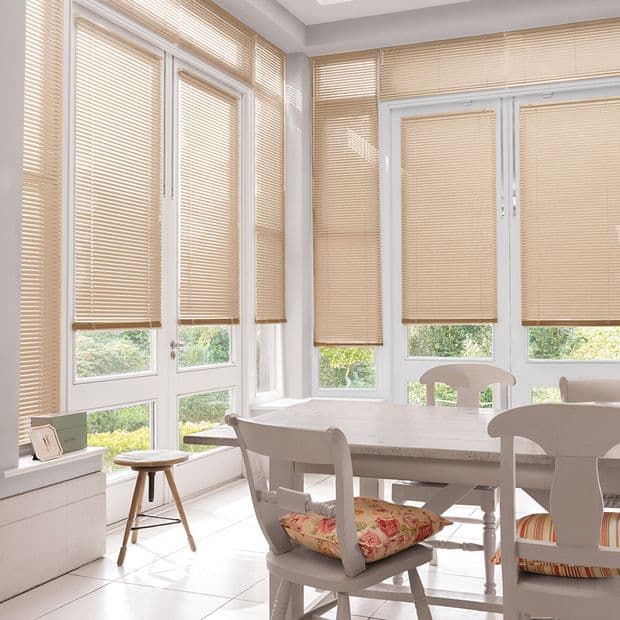 Beige horizontal Venetian blinds covering large corner windows, lowered and filtering daylight; dining table and chairs set nearby in a bright sunlit breakfast nook with a garden view.