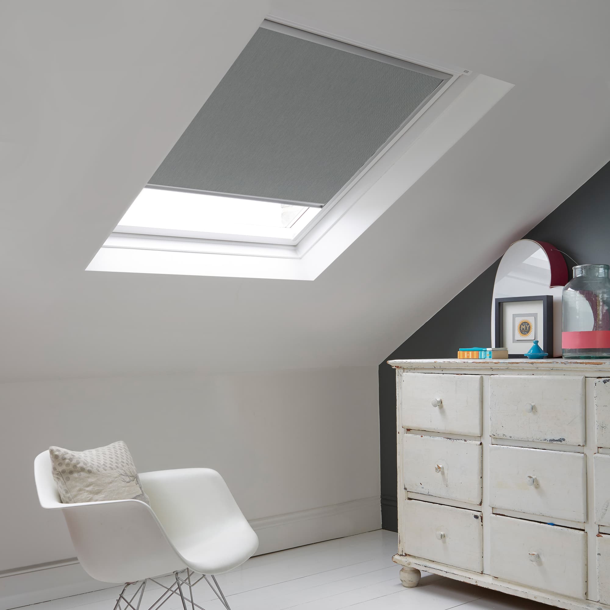 Room with white eaves wall, dark blue-grey wall to right with a shabby chic white drawer unit, a contemporary white chair, white painted floorboards and a skylight blind in mid grey.