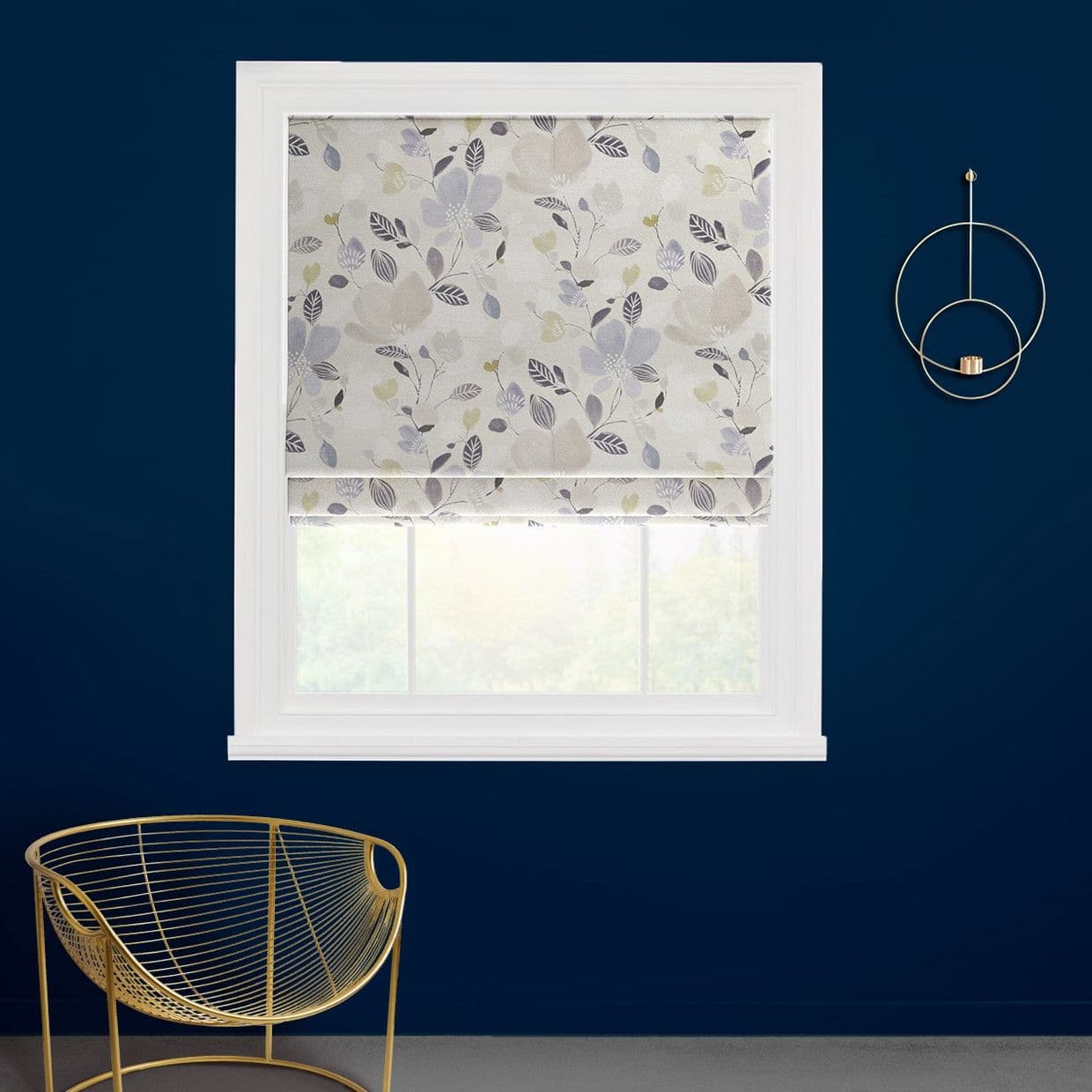 A floral-patterned Roman blind, partially lowered with soft pleats across a white-framed window, filtering daylight into a navy-painted room with a gold wire chair and circular wall sconce.