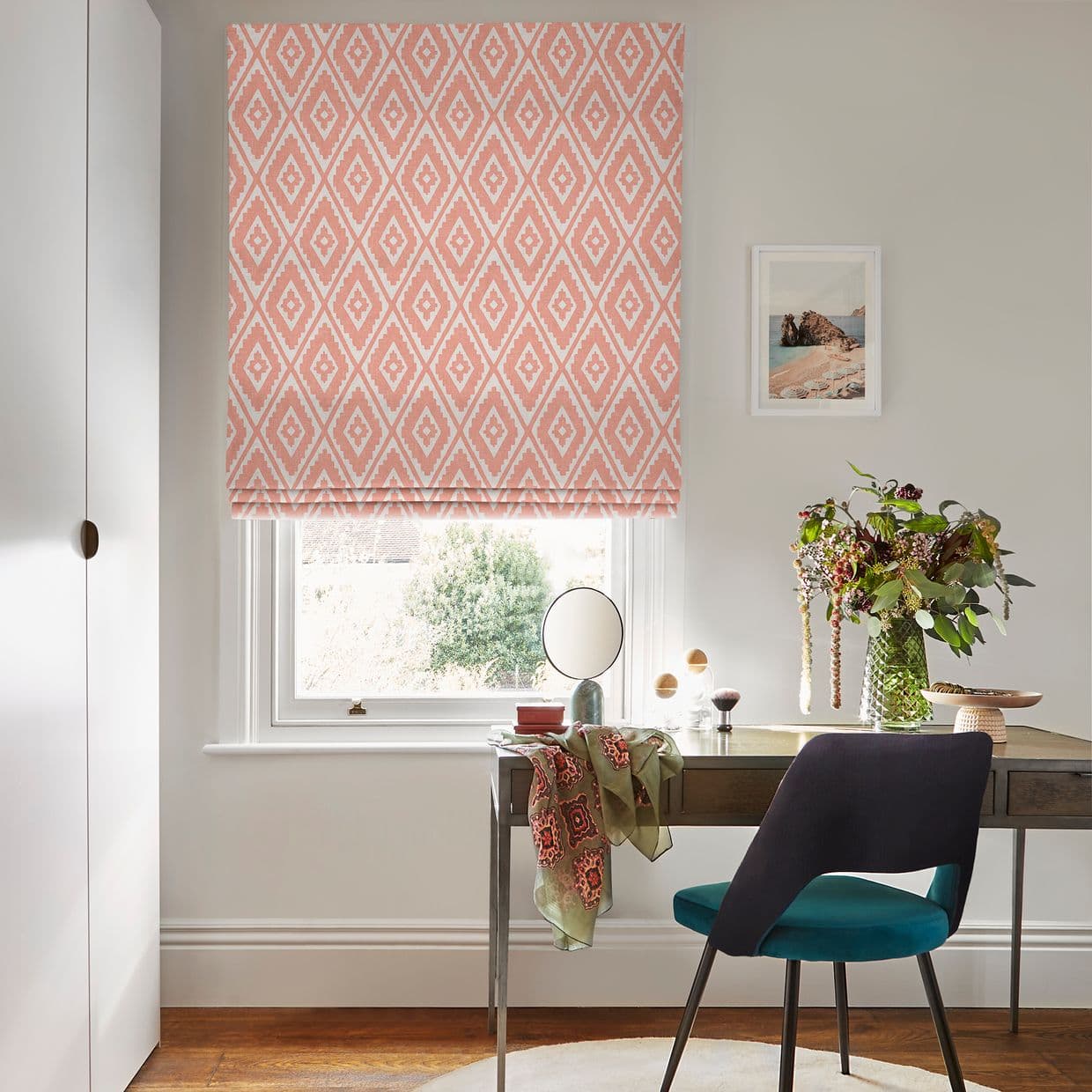Pink patterned Roman blind, fully lowered over a sash window, filtering bright natural light; simple desk with teal-upholstered chair, round mirror, vase of foliage, and a framed seaside print on a neutral wall.