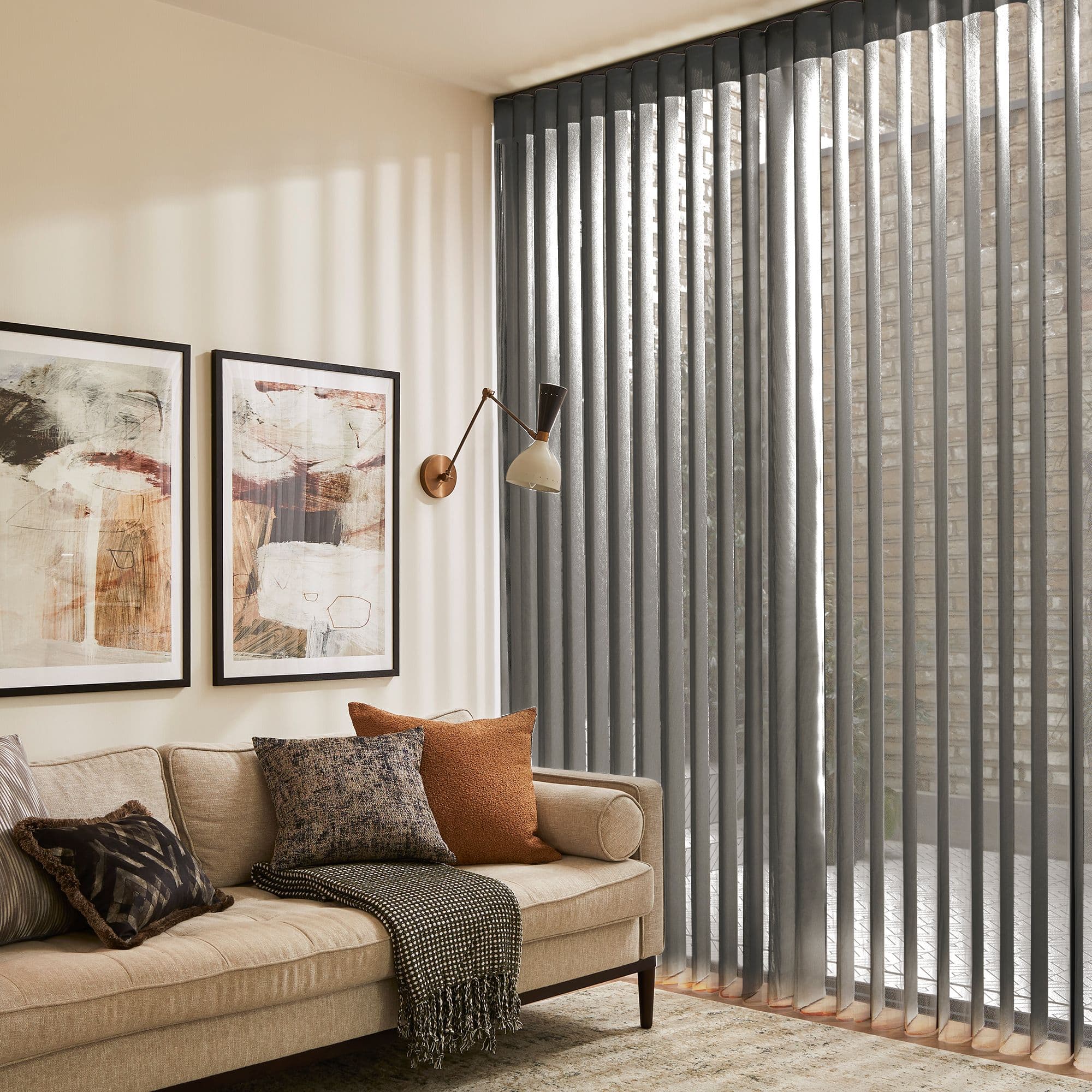 Grey floor-to-ceiling vertical blinds covering a sliding glass door, slightly angled and filtering daylight, casting striped shadows across a cozy beige living room with sofa, cushions, and abstract wall art.