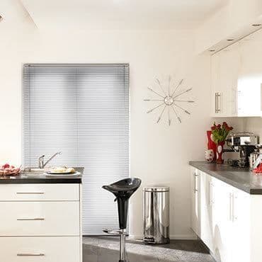 Horizontal aluminum blinds covering a narrow window, fully closed and diffusing daylight; modern white kitchen with sink, countertops, bar stool, wall clock, trash bin, vase of red tulips.