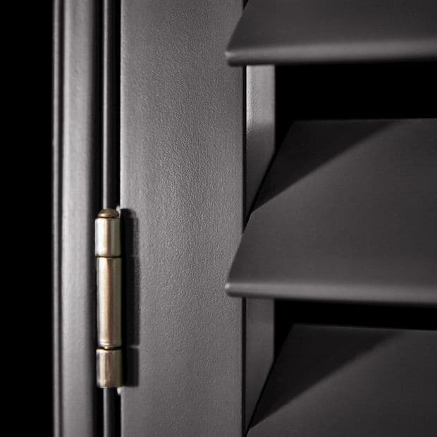 Matte dark louvered window shutters with angled horizontal slats and a brass hinge at left â€” slats closed, casting sharp shadows in a close-up interior view with low directional lighting.