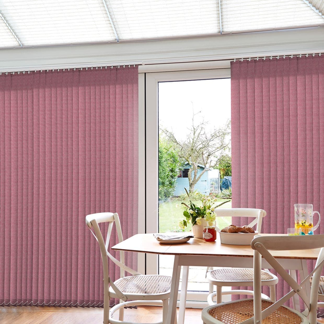 Vertical pink fabric blinds, mostly closed with a central gap, filtering bright daylight; wooden dining table with caneâ€‘seat chairs inside, framed view of a tree and garden shed outside.