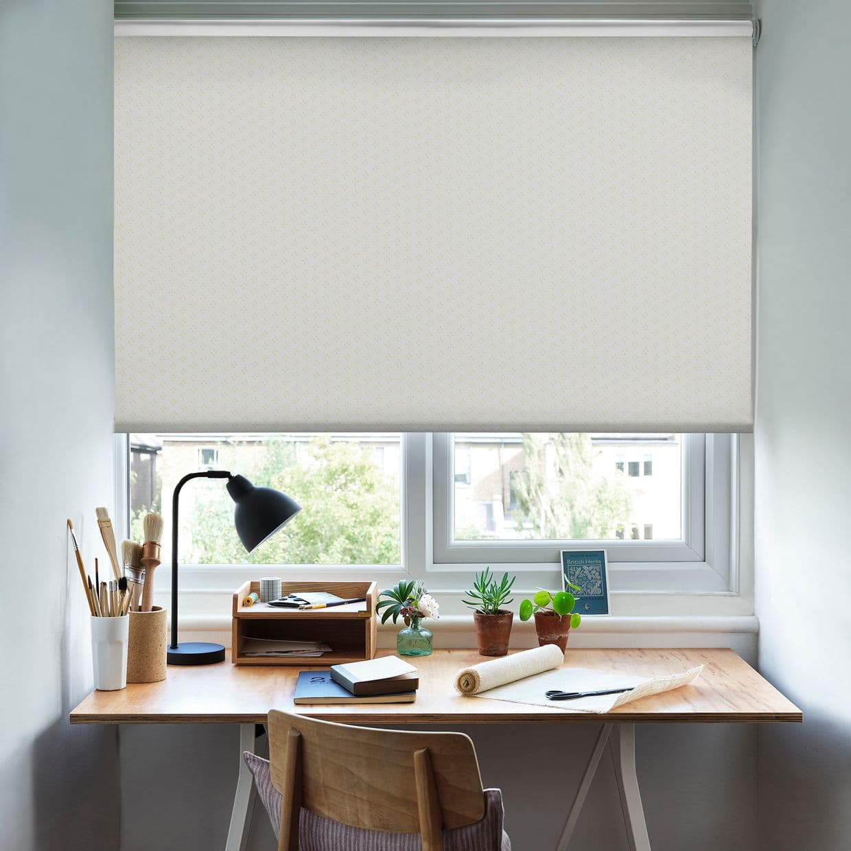 A white patterned roller blind pulled down mid-window, softly filtering daylight; below, a wooden desk with lamp, brushes, notebooks and small potted plants in a bright, tidy home workspace.