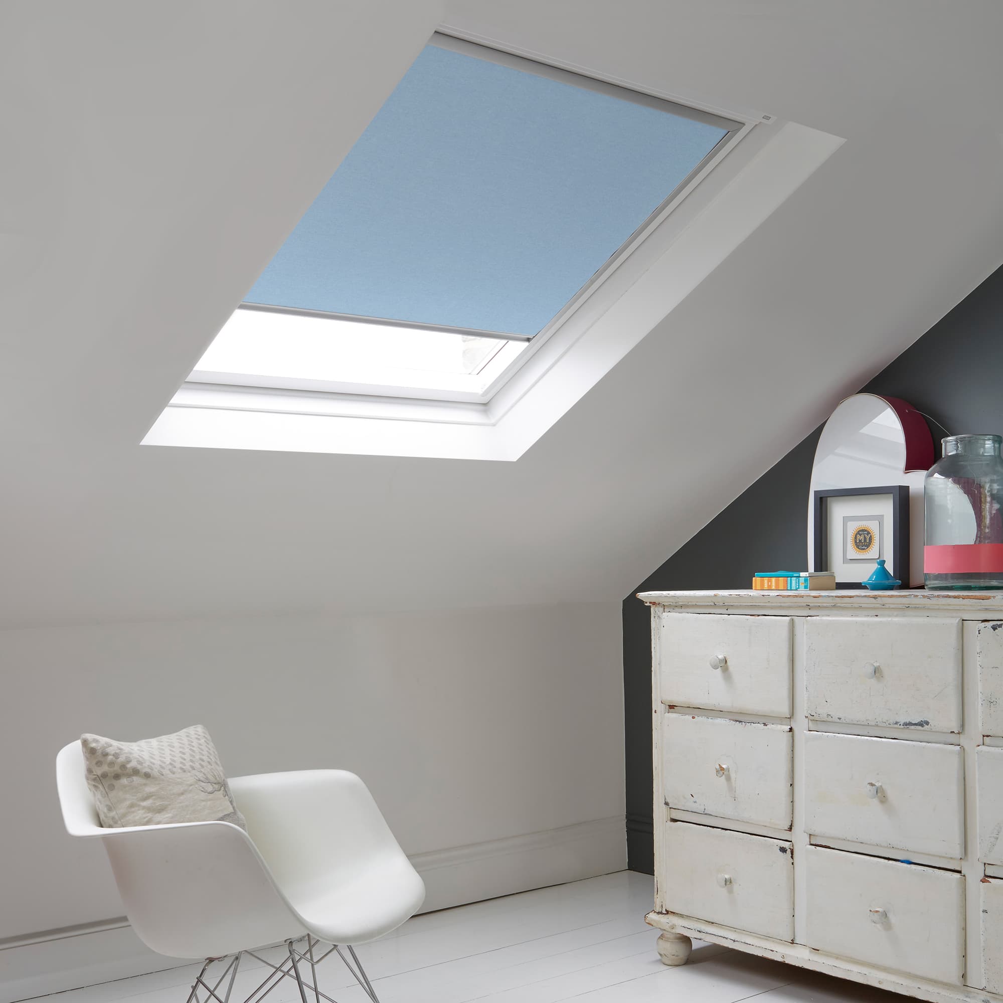 Room with white eaves wall, dark blue-grey wall to right with a shabby chic white drawer unit, a contemporary white chair, white painted floorboards and a skylight blind in pale cornflower.