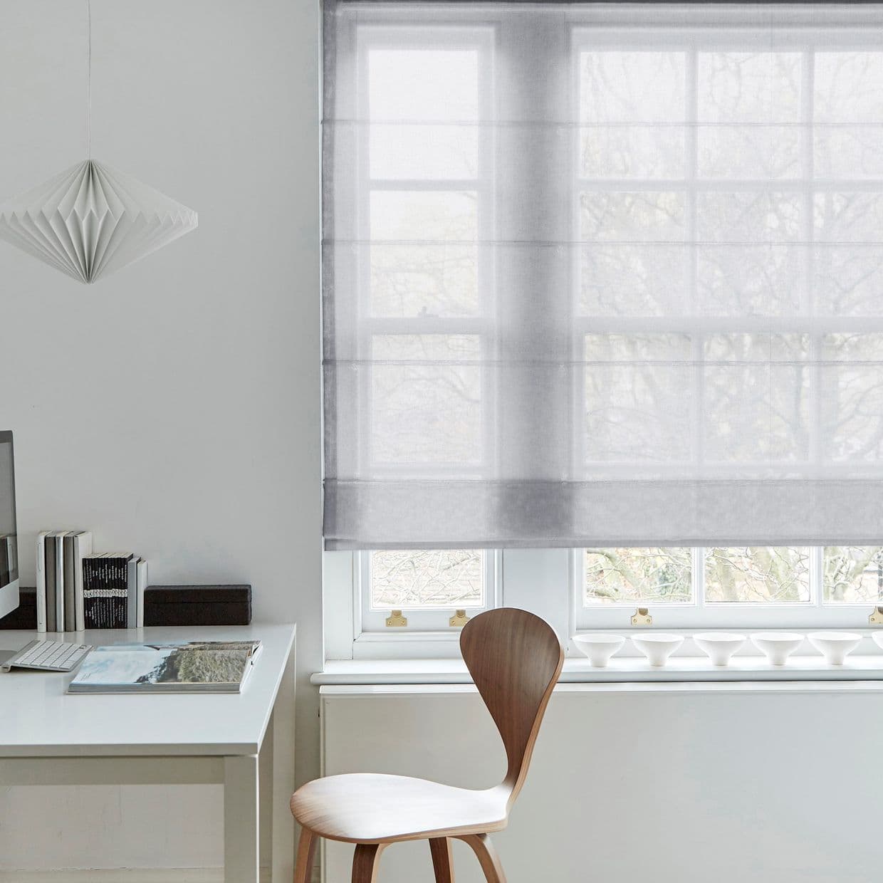 Sheer gray Roman blind lowered over a multi-pane window, softly filtering daylight; minimalist home office with white desk, wooden chair, hanging paper pendant light and a small stack of books.