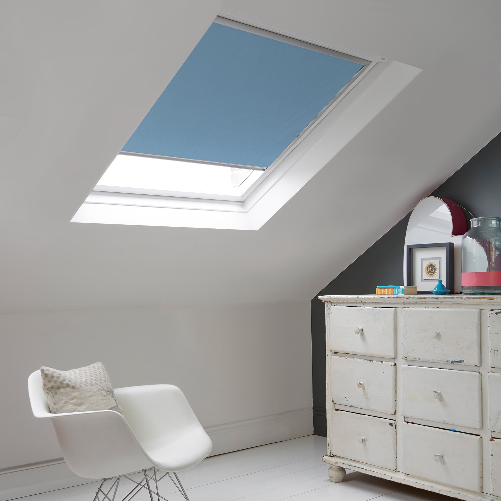 Room with white eaves wall, dark blue-grey wall to right with a shabby chic white drawer unit, a contemporary white chair, white painted floorboards and a skylight blind in pale sea blue.